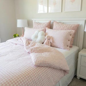 May include: A bed with a white headboard and a pink and white gingham duvet cover and pillowcases. A white cloud-shaped cushion and a pink stuffed bunny are on the bed. A white lamp and artwork are visible.
