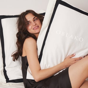 Pode incluir: Uma mulher com um vestido preto est&aacute; segurando um travesseiro branco com acabamento preto. O travesseiro tem a palavra "OBLAKO" impressa em branco.