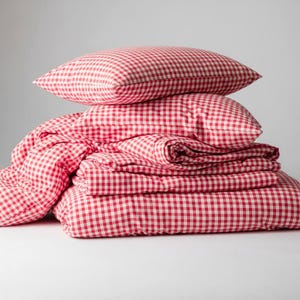 May include: A stack of red and white gingham bedding, including two pillows, a duvet cover, and a fitted sheet. The bedding is neatly folded and stacked, creating a cozy and inviting aesthetic. The classic pattern adds a touch of rustic charm.