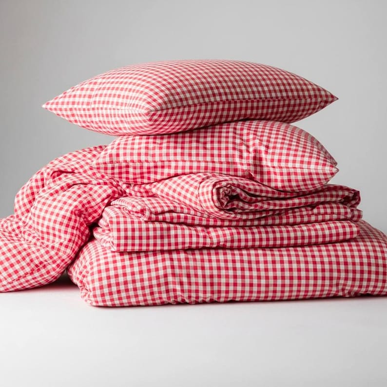 May include: A stack of red and white gingham bedding, including two pillows, a duvet cover, and a fitted sheet. The bedding is neatly folded and stacked, creating a cozy and inviting aesthetic. The classic pattern adds a touch of rustic charm.