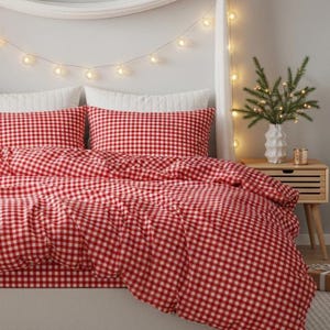 May include: A red and white gingham patterned bedding set. The set includes a duvet cover and two pillowcases. The bed is made up with white pillows and a white bed skirt. A small wooden nightstand with a vase of greenery and fairy lights is next to the bed.