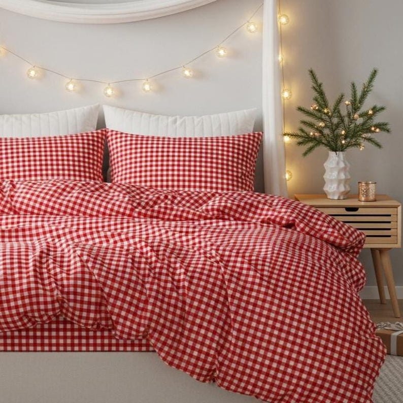 May include: A red and white gingham patterned bedding set. The set includes a duvet cover and two pillowcases. The bed is made up with white pillows and a white bed skirt. A small wooden nightstand with a vase of greenery and fairy lights is next to the bed.