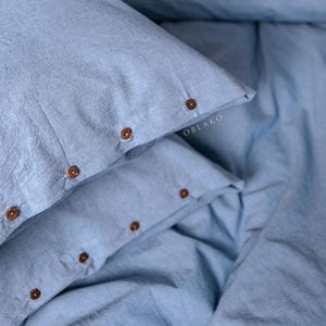 May include: A close-up of a blue linen bedding set with brown button closures. The bedding is folded and arranged in a way that shows the texture of the fabric.