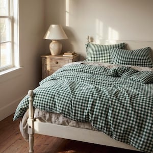 May include: A bed with a white frame and a green and white checkered duvet cover and pillowcases. A beige lamp sits on a wooden nightstand next to the bed. The room has a window and wooden floors.