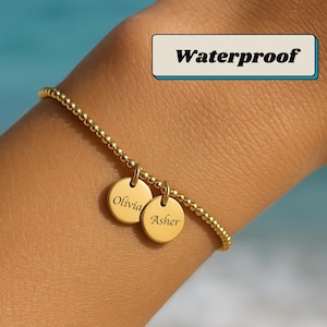Puede incluir: Pulsera dorada de cuentas con dos colgantes redondos. Un colgante dice "Olivia" y el otro "Asher". La palabra "Waterproof" está en un cuadro blanco con un contorno azul. La pulsera está en una muñeca.