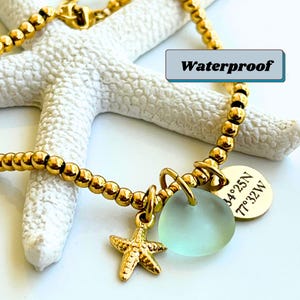 May include: Gold-colored beaded bracelet with a starfish charm, a light blue sea glass pendant, and a gold disc with coordinates. The word "Waterproof" is displayed. The bracelet is resting on a white starfish.