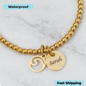 Puede incluir: Pulsera dorada con cuentas con dos colgantes. Un colgante es un colgante circular grabado con el nombre "Sarah". El otro colgante es un diseño de ola. La palabra "Waterproof" está en la imagen.