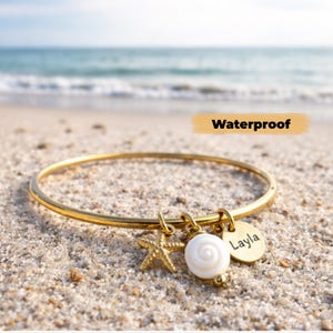 May include: A gold-colored bangle bracelet with three charms: a starfish, a white seashell, and a round tag with the name "Layla". The word "Waterproof" is written in black on a tan background. The bracelet is on a sandy beach.