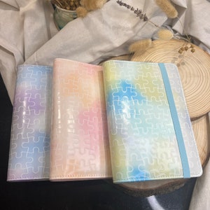 Könnte beinhalten: Drei Notizbücher in Pastellfarben mit einem weißen Puzzle-Muster. Die Notizbücher haben einen Farbverlauf von Blau, Rosa und Gelb. Jedes Notizbuch hat ein blaues Band als Lesezeichen.