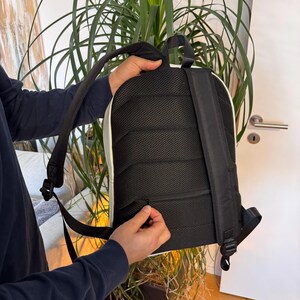 May include: Black backpack with white trim and a black mesh back panel. The backpack has adjustable shoulder straps and a zippered compartment. The person is holding the zipper of the backpack.