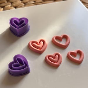 Op de afbeelding: Vijf hartvormige koekjesuitstekers, twee paars en drie roze. De uitstekers zijn dubbelwandig, waardoor een gedetailleerde hartvorm ontstaat.