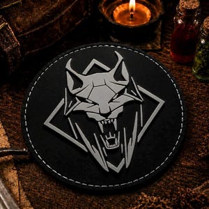 May include: A circular black patch featuring a raised, light gray stylized wolf head emblem centered within a diamond-shaped frame. The patch has a stitched border and a matte finish, displaying a sharp, aggressive design with visible fangs.