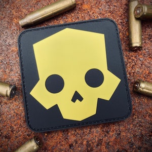 Puede incluir: Parche cuadrado negro con un diseño de calavera amarilla. Las vainas de bala de latón rodean el parche, sobre un fondo oxidado y texturizado. La calavera es de diseño geométrico.