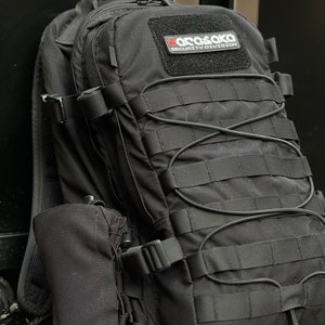 May include: Black tactical backpack with multiple compartments, straps, and a water bottle holder. Features a patch with the text "Arasaka Security Division" in red and white. The backpack has a rugged, durable design.