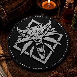 May include: A circular black patch featuring a raised, silver-colored wolf head emblem with sharp, geometric lines and an open mouth showing teeth, framed by a white stitched border, centered on a dark, textured surface with a sword nearby.