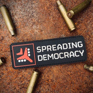 May include: A black patch with the words "SPREADING DEMOCRACY" in white lettering, and a red and white graphic. Several spent bullet casings surround the patch on a rusty, textured surface.