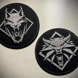 May include: Two circular black patches featuring a raised, silver-colored wolf head emblem with sharp teeth and piercing red eyes. The wolf design is set against a black background within a diamond-shaped frame, all enclosed in a circular border.
