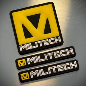 May include: Three black and yellow MILITECH patches are displayed. The largest is square with a yellow centre and black border. The word MILITECH is in white block letters. Two smaller rectangular patches also feature the MILITECH logo.