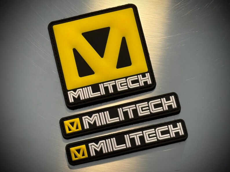 May include: Three black and yellow MILITECH patches are displayed. The largest is square with a yellow centre and black border. The word MILITECH is in white block letters. Two smaller rectangular patches also feature the MILITECH logo.