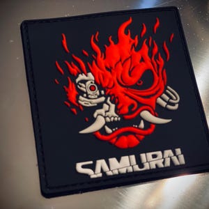 May include: A black rectangular patch with a red and white samurai mask design and flames. The word "SAMURAI" is printed in white at the bottom. The patch has a textured surface.