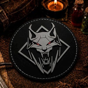 May include: A circular black patch featuring a raised, light gray geometric wolf head emblem with sharp, angular lines and piercing red eyes, set against a dark, textured background with a metallic sword and small glass vials.