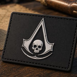 May include: A rectangular black patch featuring a silver-toned emblem of a stylized triangular crest with a skull centered within the design. The patch has a stitched border and a matte finish, presenting a clean and sharp graphic representation.