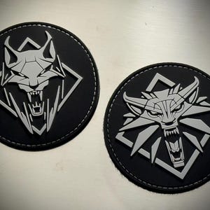 May include: Two circular black patches featuring a raised, light gray geometric wolf head design. Each wolf is depicted with an open mouth showing teeth, set against a diamond-shaped frame. The patches have a smooth, matte finish and stitched edges.