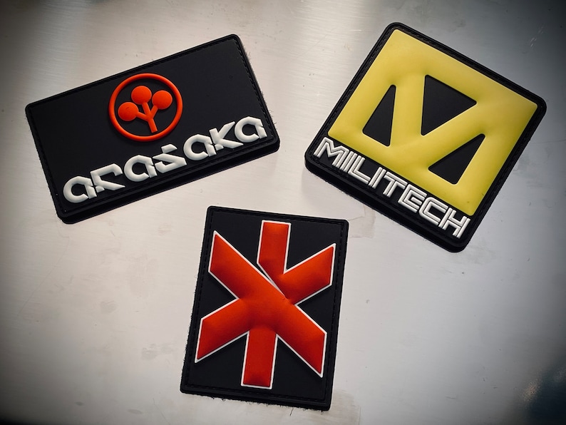 May include: Three rectangular patches with logos. One black patch has an orange logo and the word "ARASAKA" in white. Another has a yellow logo and the word "MILITECH" in white. The third has a red starburst design.