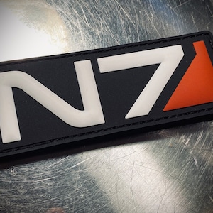 Könnte beinhalten: Ein schwarzer Patch mit weißer N7-Beschriftung und einem roten Dreieck. Der Patch ist aus einem strukturierten Material gefertigt und hat einen genähten Rand.