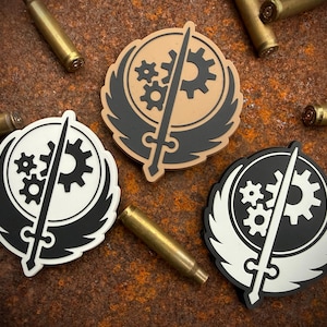 May include: Three circular patches with a sword and gear design, in black, white, and tan, are displayed on a rusty brown surface. Several brass bullet casings are scattered around the patches, adding a military or industrial theme.