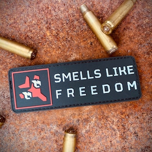 May include: A black rectangular patch with the words "SMELLS LIKE FREEDOM" in white block letters. A red and white graphic is on the left. Several spent brass bullet casings are scattered around the patch on a rusty metal surface.
