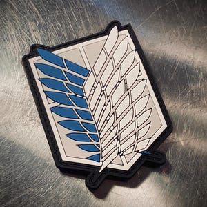 Può includere: Una toppa bianca e nera con un design ad ala blu e bianca. La toppa ha un bordo nero ed è a forma di scudo. Il testo "Wings of Freedom" è stampato sulla toppa.