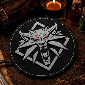 May include: A circular black patch featuring a silver-colored wolf head emblem with sharp, angular features and glowing red eyes. The wolf is centered within a diamond-shaped frame, and the entire design is stitched onto a dark, circular base.