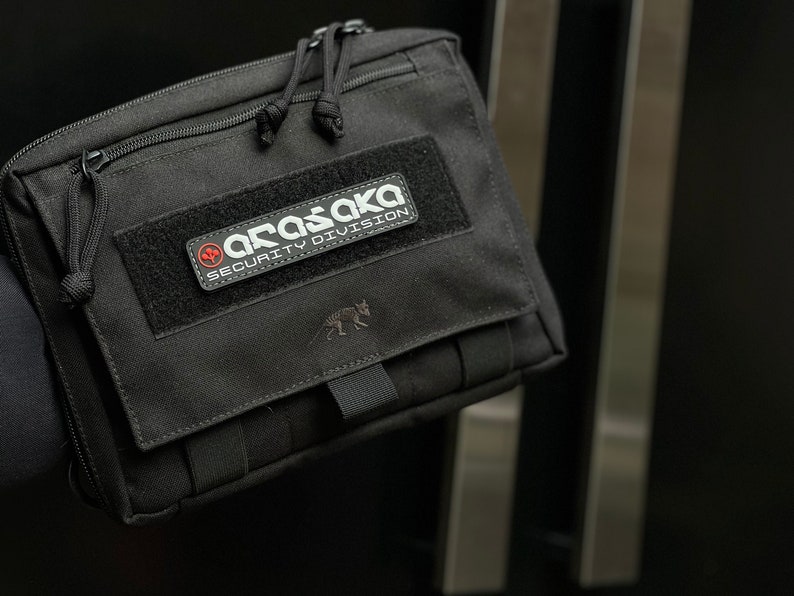 May include: Black rectangular pouch with a zippered top and a patch reading "ARASAKA SECURITY DIVISION". The pouch features a small animal silhouette and a black strap. The background is dark.