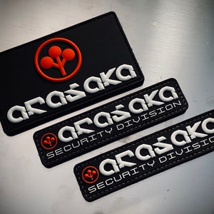 May include: Black rectangular patches with the word "ARASAKA" in white, stylized lettering. One patch has an orange logo. Two smaller patches read "ARASAKA SECURITY DIVISION".