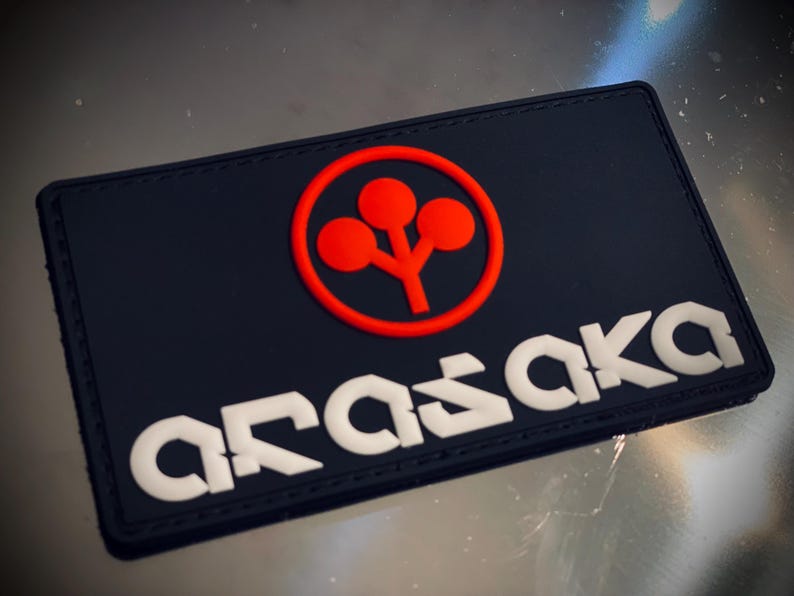 May include: A black rectangular patch featuring the white word "ARASAKA". Above the text is a red circle containing a red, stylized tree design. The patch has a textured surface.