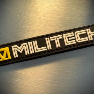 Puede incluir: Un parche rectangular negro con la palabra "MILITECH" en letras blancas. El parche tiene un cuadrado amarillo con una "M" blanca en el lado izquierdo. El parche está sobre una superficie metálica reflectante.