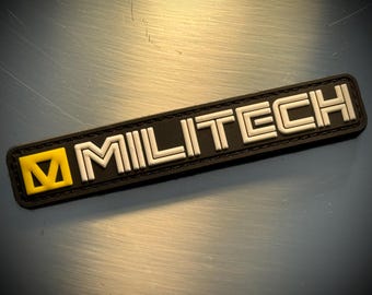 May include: A black rectangular patch with the word "MILITECH" in white, block letters. The patch has a yellow square with a white "M" on the left side. The patch is on a reflective, metallic surface.