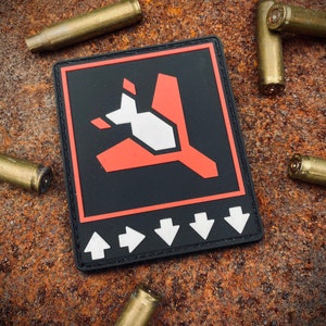 May include: A black and red patch with a stylized graphic of a plane or bird, surrounded by white arrows. The patch is on a rusty metal surface, with several brass bullet casings scattered around it.