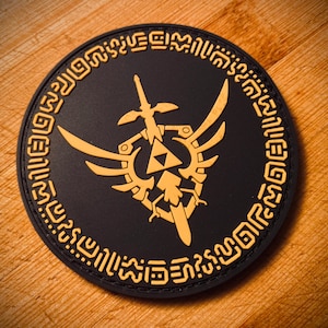 May include: A round, black patch featuring a gold design of a sword, wings, and a triangle, surrounded by a gold border with stylized text. The patch is likely a collectible or emblem.
