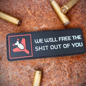 May include: Black rectangular patch with red and white graphic and the text "WE WILL FREE THE SHIT OUT OF YOU." Several brass bullet casings are scattered around the patch on a rusty metal surface.