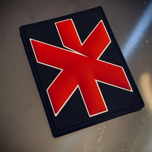 May include: A rectangular patch featuring a navy blue base and a prominent red graphic, outlined in white. The patch has a black border. The design is likely an emblem or logo.
