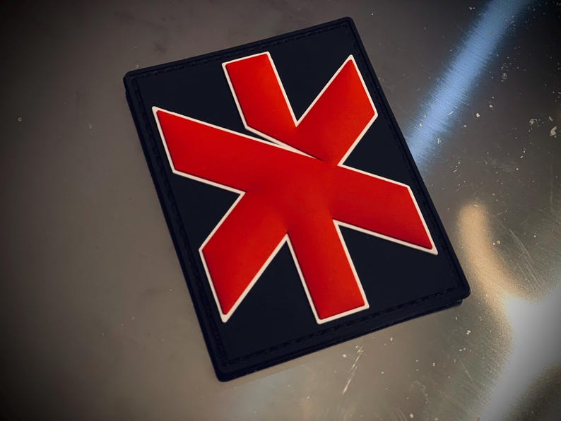 May include: A rectangular patch featuring a navy blue base and a prominent red graphic, outlined in white. The patch has a black border. The design is likely an emblem or logo.