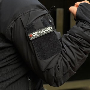 May include: Black tactical jacket featuring a patch with the text "ARASAKA SECURITY DIVISION". The jacket has a textured pattern on the upper arm and a velcro patch for attaching the security division patch.