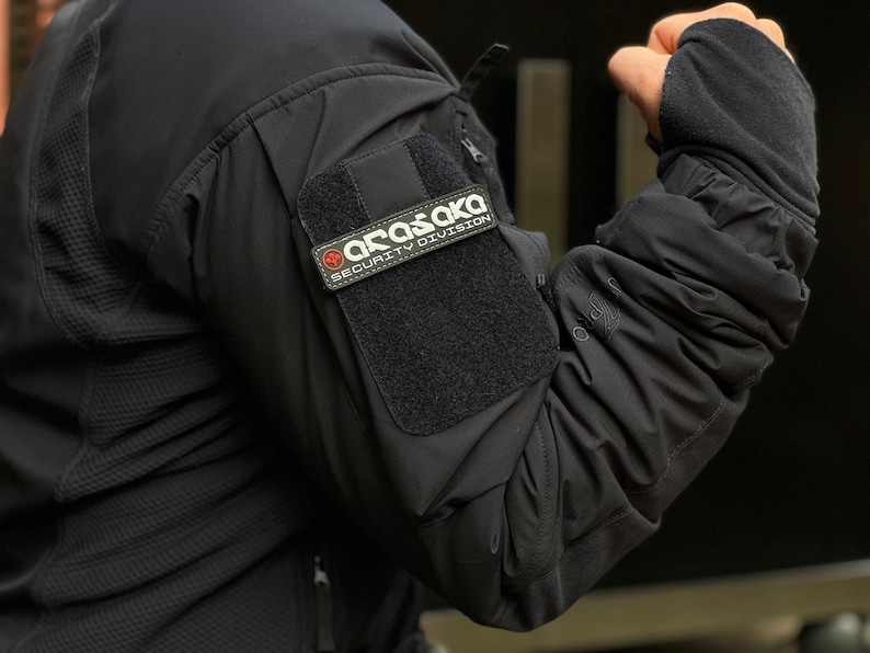 May include: Black tactical jacket featuring a patch with the text "ARASAKA SECURITY DIVISION". The jacket has a textured pattern on the upper arm and a velcro patch for attaching the security division patch.