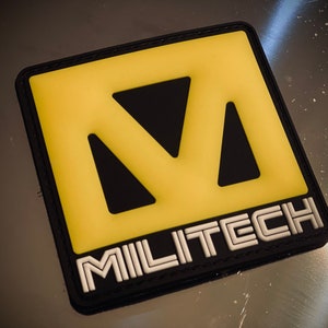 May include: A square patch with a yellow and black design. The yellow section features a stylized letter "M" with cut-out triangles. The word "MILITECH" is in white, outlined in black, at the bottom of the patch. The patch has a black border.