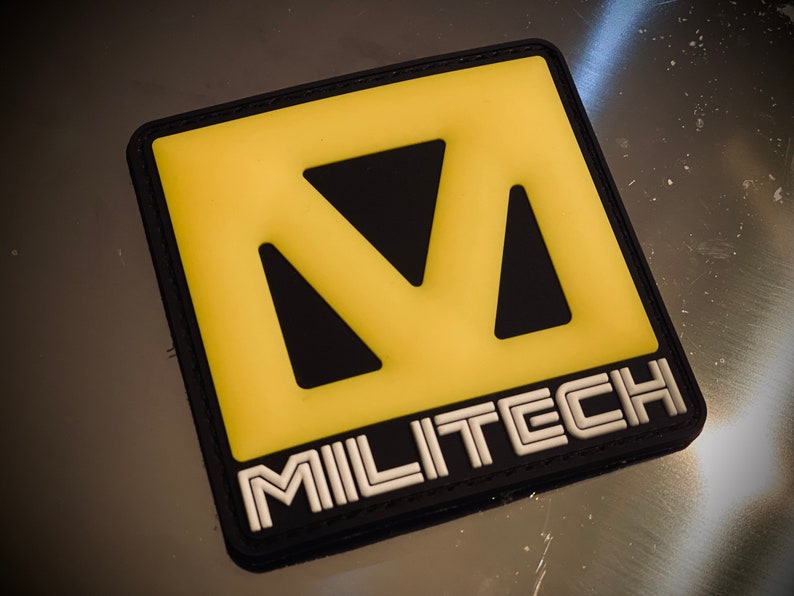 May include: Square patch with a yellow and black design. The yellow section features a stylized letter "M" with cut-out shapes. The word "MILITECH" is displayed in white, outlined in black, at the bottom of the patch. The patch has a black border.