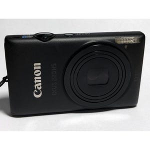 Könnte beinhalten: Schwarze Canon IXUS 220 HS Digitalkamera. Die Kamera hat ein Objektiv, einen Blitz und verschiedene Knöpfe. Das Canon-Logo und der Modellname sind weiß auf der Vorderseite aufgedruckt. Ein schwarzer Riemen ist befestigt.