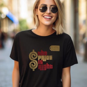 May include: A black short-sleeved t-shirt featuring a gold dollar sign and the text "the PRICE is right" in a mix of gold and red glittery lettering. A small gold rectangular price tag graphic is positioned above the text on the chest.