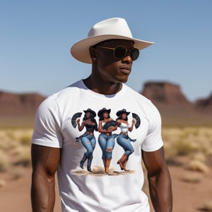 Op de afbeelding: Wit T-shirt met een afbeelding van drie vrouwen met cowboyhoeden, die waaiers vasthouden en een spijkerbroek en laarzen dragen. De afbeelding is geplaatst tegen een woestijnachtergrond. De persoon op de afbeelding draagt een witte hoed en een zonnebril.